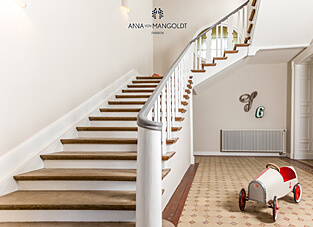 Ein helles, weitläufiges Treppenhaus mit einer geschwungenen Holztreppe. Die Wände sind in der cremigen Weißfarbe "Downton Abbey" von ANNA VON MANGOLDT FARBEN gestrichen. Die Treppe hat weiße Setzstufen und Holztrittflächen. Der Boden ist mit gemusterten, historischen Fliesen in Beige und Braun belegt. Im unteren Bereich steht ein altmodisches Tretauto in Weiß und Rot.