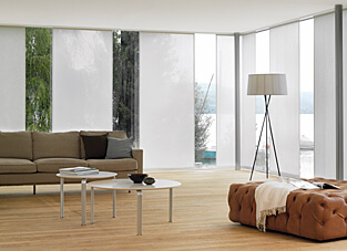 Ausschnitt eines modernen Wohnbereichs mit großer Fensterfront über Eck. An der Fensterfront weiße Flächenvorhänge an mehrläufigen Einnut-Profilen.