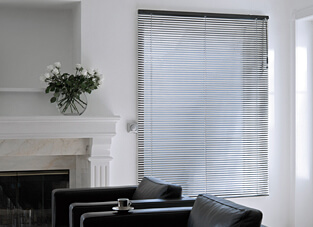 Ausschnitt eines Wohnbereichs. Weiße Wände. Rechts en Fenster mit Jalousie mit Aluminium-Lamellen und Stabbedienung. Links weißer Kamin mit schwarzen Sesseln davor.