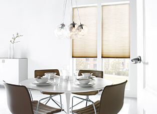Moderner Essbereich mit rundem weißen Tisch und vier braunen Stühlen darum. Gedeckt mit weißem Geschirr. An den fenstern beige Plissees. mit Elektroantrieb.
