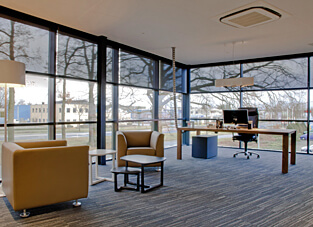 Modernes Büro mit dunklem Fußboden, links eine Sitzgruppe aus zwei gelb orangenen Sesseln und zwei kleinen dunklen Beistelltischen. Im Hintergrund ein großer Schreibtisch aus Holz mit dunklem Bürostuhl. Umgeben von einer raumhohen Fensterfront über Eck versehen mit Rollos aus Screen-Stoff.