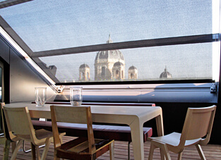Essbereich mit großem Holztisch umgeben von drei Stühle und deiner roten Sitzbank. Im Hintergrund ein großes schräges Dachfenster mit Skylight-System in Rolloform.