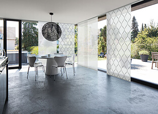 Ein moderner, heller Essbereich mit Betongussboden und großen Glasfronten zum Garten. Die Fenster sind mit einem Flächenvorhangsystem versehen, das verschiedene Designs kombiniert (Designmix). Es sind transparente Paneele mit einem weißen Rauten- oder Spitzbogenmuster sowie ein unifarbenes, transparentes Paneel hinter einer runden, dunklen Hängelampe zu sehen. Ein weißer, runder Tisch mit grauen Stühlen steht im Raum.