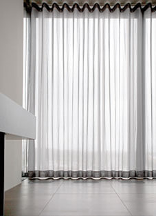 Eine helle Fensterfront, die fast vollständig von einer halbtransparenten Gardine (Store) von Silent Gliss bedeckt ist. Der Stoff fällt in gleichmäßigen, vertikalen Falten, erzeugt durch das Wave-Wellenband und ist an den oberen und unteren Kanten mit einem dunklen Wellensaum versehen. Die Gardine ist schwer entflammbar und lässt viel Licht in den Raum mit dunklem Fliesenboden.
