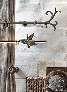 Designbeispiel von zwei Gardinenstangen mit Ringe auf heller Wand in Holzoptik montiert. Oben Messing brüniert mit runder Halterung und Endstück Hamlet. Unten Messing mit grün melierter Halterung in Blattform.