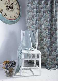 in romantisch gestalteter Raum im Landhausstil mit hellen Holzböden und einer rustikalen, türkisfarbenen Wanduhr. Ein Dekorationsstoff von Art of the Loom hängt als Vorhang und weist ein florales, wiederkehrendes Muster in gedämpften Blau-, Grün- und Grautönen auf. Auf einem weißen Stuhl liegt ein hellblauer Überwurf. Eine Vase mit dunkelblauen Hortensien steht links am Boden.