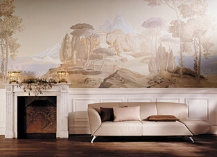 Ein elegantes Wohnzimmer mit weißer Holztäfelung und einem weißen Marmorkamin. Die Wand ist mit einem großformatigen Wandbild (Mural) versehen, das eine antike, mediterrane Landschaft in sanften Pastell- und Beigetönen zeigt. Das Wandbild hat eine spezielle Struktur, da es auf einem gecrushten Vliesträger gedruckt ist, der die Oberfläche texturiert. Davor steht ein modernes, beigefarbenes Designersofa.