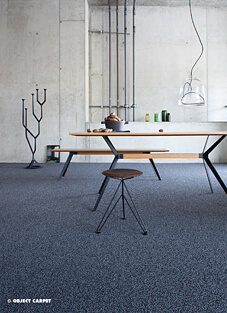 Ein modern gestalteter Essbereich im Industrial-Stil mit Sichtbetonwänden und freiliegenden Rohren. Der Boden ist mit einem getufteten Objektschlingen-Teppichfliesen in dunkelblau-grauer Kieselsteinoptik ausgelegt. Ein Holztisch mit schwarzen Metallbeinen und eine moderne Hocker-Skulptur stehen auf dem Teppich.