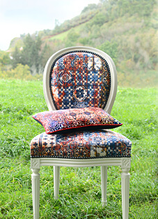 Ein historisch anmutender, weiß lackierter Stuhl im Freien, platziert auf einer grünen Wiese vor einer hügeligen Landschaft. Der Stuhl ist mit dem Stoff von Jean Paul Gaultier im Landhausstil bezogen. Der Stoff weist ein patchworkartiges, farbenfrohes Mosaikmuster in Rostrot, Blau und Beige auf. Ein passendes, kleines Kissen liegt auf dem Sitz.