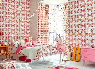 Ein helles und mädchenhaftes Kinderzimmer, das vollständig auf ein Schmetterlingsmotiv abgestimmt ist. Die Wände sind mit einer Tapete mit großen, rosa und orangefarbenen Schmetterlingen bedeckt, ergänzt durch einen Bereich mit einem kleineren, gesprenkelten Muster. Ein weißes Metallbett ist mit einer Decke mit Schmetterlingsmuster und pinken Kissen ausgestattet. Vor dem Fenster hängen passende Schmetterlings-Vorhänge. Die Einrichtung umfasst eine pinke Kommode, einen orangenen Nachttisch und einen großen, orangenen Buchstaben 'B' auf dem Boden.