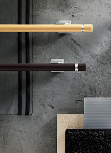 Designbeispiel mit zwei Gardinenstangen, montiert auf einer grauen Betonwand mit silbernen Halter. Oben helles Holz, unten Wenge Farben. Beide mit silbernen Ring am Endstück.