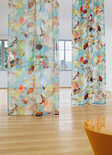 Ein heller Raum mit Holzboden und weißen Wänden. Zwei hohe, halbtransparente Dekorationsstoffe von Création Baumann hängen im Raum. Der Stoff hat ein romantisches, florales Muster aus großen, überlappenden Formen in Pastellfarben, darunter Blau, Rosa, Gelb und Grün, das dem Stil Landhaus ähnelt.