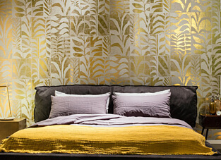 Ein Schlafzimmer mit einem großen, dunklen Bett, das mit einem senfgelben Plaid und lila Kissen dekoriert ist. Die Wand hinter dem Bett ist mit einer glänzenden Vliestapete versehen, die ein großflächiges, geometrisches Palmenblattmuster in Gold auf einem hellen Hintergrund zeigt.