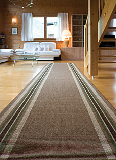 Ein Sisalläufer in bewährter Mellau-Qualität liegt auf einem hellen Holzboden in einem rustikalen, holzgetäfelten Wohnzimmer. Der Läufer hat eine braune Grundfarbe und wird von schmalen Streifen in Beige, Weiß und verschiedenen Grüntönen gesäumt. Im Hintergrund sind eine weiße Couch und eine Holztreppe sichtbar.