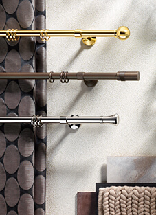 Designbeispiel von drei Gardinenstangen mit Ringen daran, montiert auf weißer Wand. Von oben nach Unten: Golden mit goldener Kugel als Endstück, mittig brüniert mit geradem Endstück, unten Silber mit konisch auslaufendem Endstück.