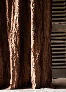 Nahaufnahme eines schweren, braunen Vorhangstoffs mit einer tiefen, geknitterten Struktur. Der Stoff besitzt einen brillanten Metall-Lüster (Glanz), der die Falten und die Textur des Materials hervorhebt. Rechts ist eine Wand mit horizontalen Lamellen in Dunkelgrau sichtbar.