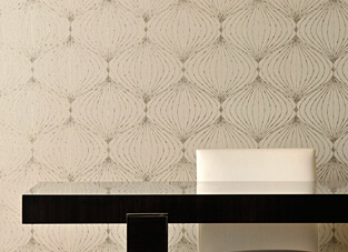 Nahaufnahme einer Wandbekleidung mit einem eleganten, grafischen Muster in Creme und Beige. Das Muster besteht aus sich überlappenden, geschwungenen Rautenformen im Stil des Art déco, die mit einem feinen Metall-Druck akzentuiert sind. Im Vordergrund verläuft die glänzende, dunkle Oberfläche eines Tisches.