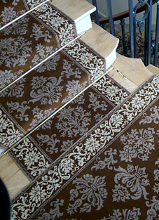 Blick von oben auf eine helle Holztreppe, deren Stufen mit einem dunkelbraunen Treppenläufer im Stil "Venise" belegt sind. Der gewebte Woll-Läufer zeigt ein opulentes, hellbraun-beiges Muster mit großen floralen Ornamenten und einer dekorativen Bordüre in Kontrastfarben. Der Läufer ist mit Teppichstangen befestigt.