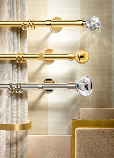 Designbeispiel von drei Gardinenstangen mit Ringen darauf, montiert auf heller Wand. von oben nach Unten: golden mit Svarowski-Kristallkugel als Endstück, mittig golden mit goldener Kugel als Endstück und unten silbern mit Svarowski-Kristall als Endstück.