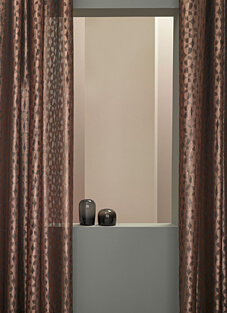 Ein elegantes Fensterarrangement, das von einem klassischen Dekorationsstoff von NYA Nordiska eingerahmt wird. Der Stoff ist in einem glänzenden Kupferton gehalten und weist ein subtiles, geometrisches Rauten- oder Wabenmuster auf. Auf der Fensterbank vor einer beigefarbenen Wand stehen zwei kleine, graue Glasvasen.