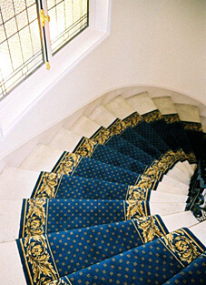 Blick von oben auf eine geschwungene, weiße Marmortreppe. Die Stufen sind mit einem klassischen, dunkelblauen Treppenläufer im Stil von "Louvres" belegt. Der gewebte Woll-Läufer ist mit einem kleinen, goldenen Punktmuster und einer breiten, goldenen Bordüre mit aufwendigen Barock- oder Akanthus-Ornamenten verziert.