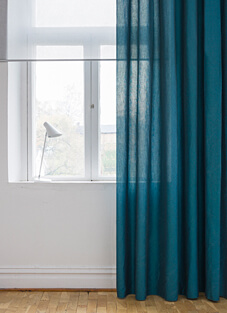 Ein heller Raum mit einem Fensterrahmen. Ein halbtransparenter Dekorationsstoff von Almedahls in einem tiefen Petrolblau hängt an einer dünnen Stange vor der rechten Fensterhälfte. Der Stoff lässt das Tageslicht gedämpft durchscheinen. Auf der weißen Fensterbank steht eine kleine, moderne Leselampe.