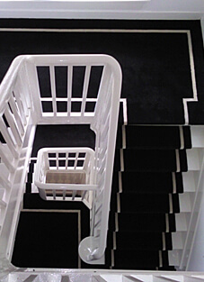 Ansicht eines Treppenhauses innen on oben nach Unten. Die Stufen und Etagen sind mit einem schwarz weißen Bordürenläufer versehen. Das Geländer und die Wände sind weiß gehalten.