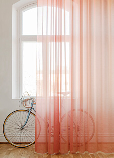 Ein heller Raum mit einem Fenster mit Rundbogen. Ein Dekorationsstoff von Almedahls, der halbtransparent ist, hängt vor dem Fenster. Der Stoff ist in einem warmen Lachs- oder Pfirsichton gehalten und erzeugt einen sanften Filtereffekt. Im Vordergrund steht ein altmodisches Rennrad, dessen Umrisse durch den Stoff sichtbar sind.