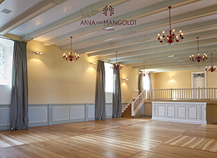 Ein großer, heller Festsaal mit Holzboden, weiß gestrichenen Deckenbalken und einer kleinen Holzempore mit weißer Balustrade. Die Wände sind in der hellen, warmen Gelb-Beige-Farbe "Sarastro" von ANNA VON MANGOLDT FARBEN gestrichen. Die untere Wandpartie ist mit einer grau-blauen Täfelung versehen. Mehrere rote Kronleuchter und graue Vorhänge ergänzen die festliche Gestaltung.