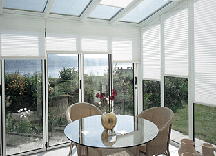 Gläserner Wintergarten mit Ausblick auf Natur. Mittig gläserner runder Tisch mit zwei geflochtenen Sesseln. An den Glasscheiben weiße Plissees.