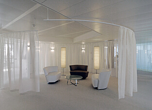 Sitzlounge in weißer Umgebung bestehend aus zwei weißen Sesseln, mittig ein brauner Sessel und einem kleinen Tisch. im Hintergrund zwei Stehlampen. Abgegrenzt wird die Lounge durch ein gebogenes Raumtrenn-System mit naturweißen Vorhängen.