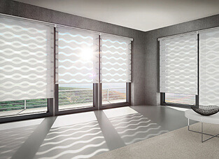 Ein moderner Raum mit grauem Boden und Betonwänden und einer breiten Fensterfront mit Meerblick. Die Fenster sind mit Wave-Doppelrollos ausgestattet, deren Stoffbahnen ein durchgehendes, wellenförmiges Muster in Weiß und Hellgrau bilden. Das durchfallende Sonnenlicht wirft das Wellenmuster als dynamische Schatten auf den Boden.