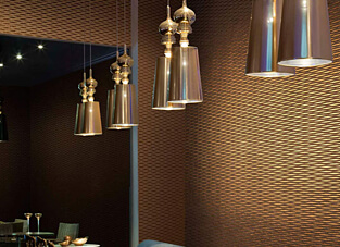 Innenansicht eines eleganten, modern beleuchteten Raumes. Die Wände sind mit einer Vliestapete in einem satten Bronze- oder Goldbraun verkleidet. Die Tapete weist eine feine, metallische Prägung in einem durchgehenden Wabenmuster auf. Mehrere moderne, hängende Pendelleuchten mit glänzenden, bronzefarbenen Schirmen hängen im Vordergrund und spiegeln sich in einer dunklen Wand.