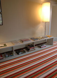 Ein heller Raum mit einer niedrigen weißen Ablage, auf der Zeitschriften liegen. Der Boden ist mit einem modernen Schurwollteppichboden ausgelegt, der ein auffälliges, gestreiftes Muster in den Farben Rot, Orange, Grün und Weiß zeigt. Im Hintergrund spendet eine hohe Stehlampe mit einem hellen Schirm warmes Licht.