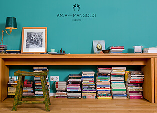 Eine Wand ist in der leuchtenden, tiefen Türkisfarbe "Esmeralda" von ANNA VON MANGOLDT FARBEN gestrichen. Davor steht ein niedriger, langer Holztisch, dessen Ablagefläche mit Büchern und einer Lampe dekoriert ist. Unter dem Tisch stapeln sich weitere Bücher in einer chaotischen, aber organisierten Weise. Links steht ein kleiner, grün lasierter Hocker.