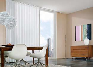 Gemütliches Esszimmer mit dunklem Holztisch und vier weißen Stühlen daran. Darüber weiße Lampen. Rechts Holzkommode und ein Bild darüber. Im Hintergrund bodentiefe Fenster mit weißem Vorhang an Elektrozug-System.