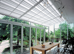 Gläserner Wintergarten, mittig großer Holztisch und Sonnenschutzsystem in Raffvorhangform.