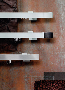 Designbeispiel von drei Gardinenschienen geradlinig gebürsteter Edelstahl, montiert auf einer Kupfer melierten Wand mit kleinen Quadratischen haltern. Oben mit drei Gleitern und konisch auslaufendem Ende, mittig mit drei Gleiter und drei Gardinenhaken und schwarzem lederner Schlaufe als Endstück, unten mit zwei Gleitern und zwei Gardinenhaken mit geradem Ende.