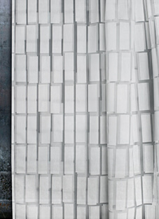 Nahaufnahme eines weißen Ausbrenner-Dekorationsstoffs von Tisca Tiara. Der halbtransparente Stoff zeigt ein modernes, grafisches und geometrisches Rechteckmuster. Der Stoff hängt vor einem hellen Fenster und die Konturen sind leicht verschwommen. Links ist eine dunkle Wandstruktur sichtbar.