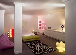 Ein modernes Schlafzimmer in Weiß mit Designer-Lampen und leuchtenden Akzenten. Der Raum ist durch klassische, weiße Zierleisten am Deckenansatz sowie durch hohe, weiße Sockelleisten an den Wänden und um die Säulen herum stilvoll eingefasst.