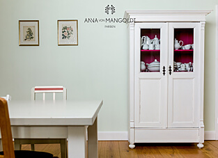 Ein heller Essbereich mit einem weißen Holztisch und einem weiß gestrichenen Schrank. Die Wand ist in der blassen, kühlen Grüntonfarbe "Ceylon" von ANNA VON MANGOLDT FARBEN gestrichen. Der Schrank hat Glastüren, hinter denen das Innere in einem leuchtenden Pink gestrichen ist und weißes Porzellan aufbewahrt wird. Zwei gerahmte botanische Drucke hängen an der Wand.