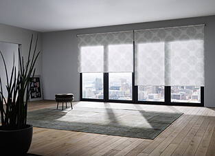 Ein modernes Loft-Zimmer mit Holzfußboden und grauen Wänden, das einen weiten Blick auf eine Stadt-Skyline freigibt. Zwei große Fenster sind mit einer Rollo-Doppelanlage versehen, deren weißer Stoff ein hellgraues, ornamentales Muster aufweist und das Licht sanft filtert. Im Vordergrund liegen ein großer, grauer Teppich und eine Topfpflanze.