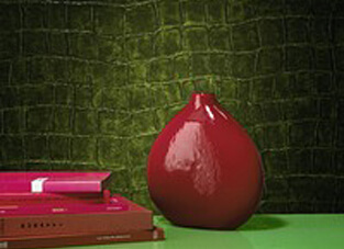 Nahaufnahme einer Oberfläche, die mit der Elitis Tapete "Croco" in Reptil-Optik tapeziert ist. Die Tapete in dunklem, glänzendem Olivgrün hat eine ausgeprägte, dreidimensionale Krokodilhautstruktur und ist modern und schwer entflammbar. Im Vordergrund stehen eine glänzende, rote Keramikvase und ein Stapel pink-roter Bücher.