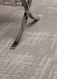 Nahaufnahme eines Bodens mit einer Teppichfliese in luxuriöser, außergewöhnlicher Tipshear-Optik. Im Vordergrund ruht das glänzende, verchromte Metallgestell eines modernen Sitzmöbels auf dem Teppich.