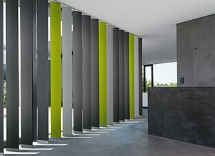 Ein moderner Raum im Industrial-Stil mit Betonwänden und grauen Böden. Eine Reihe von extra breiten (XXL) Vertikallamellen dient als Raumteiler und zeigt einen dynamischen Farbmix aus verschiedenen Grautönen und leuchtenden, limettengrünen Akzenten.
