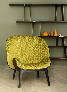 Ein moderner Loungesessel mit schwarzem Holzgestell vor einer olivgrünen Wand. Der Sessel ist mit einem Polsterstoff von Lelièvre bezogen, einem strapazierfähigen Velours in leuchtendem Olivgrün. Der klassische Stoff weist ein dichtes, geometrisches Kettenmuster auf. Im Hintergrund ist ein modernes, dunkles Regal mit dekorativen Objekten sichtbar.