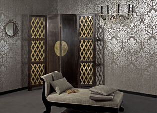 in elegant inszenierter Raum mit dunklem Boden. Die Wände sind mit einer Omexco Vliestapete in einem klassischen Ornamentmuster (Damast-Stil) tapeziert, das in metallisch glänzendem Silber und Weiß auf einem grauen Hintergrund gehalten ist. Davor stehen eine geschwungene Chaiselongue in Grau und ein dunkler Paravent mit goldenen Rauten-Einsätzen.