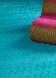 Nahaufnahme eines gewebten Vinyl-Bodenbelags von Bolon aus der Kollektion Missoni. Der langlebige und pflegeleichte Bodenbelag ist in einem leuchtenden Türkiston gehalten und weist ein subtiles, wellenförmiges Muster auf. Im oberen rechten Bereich ist die schräg angeschnittene Kante eines modernen Möbels oder Stuhls in Pink und Braun sichtbar.