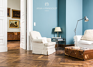 Ein Wohnraum in einem Altbau mit dunklem Fischgrätparkett und weißen, hohen Flügeltüren. Die Wand im Wohnbereich ist in der hellen, staubigen Blaufarbe "Favory" von ANNA VON MANGOLDT FARBEN gestrichen. Auf dem Boden steht ein weißer, bezogener Sessel, ein Beistelltisch mit Lampe und ein alter Lederkoffer als Couchtisch. Durch die geöffnete Tür sind gerahmte, klassische Ölgemälde in einem Nebenraum sichtbar.
