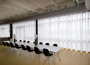 großer weißer Konferenztisch umgeben von 16 dunklen Stühlen und zwei weißen Orchideen darauf. gelber gefliester Boden, industrielle weiße Decke und lange Fensterfront mit weißen Wave XL Vorhängen.